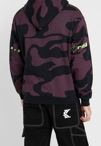 Černá mikina s kapucí, s kontrastními purpurovými svislými pruhy a vzorem maskáčů. Obsahuje kapuci a zelené textové akcenty na rukávu.