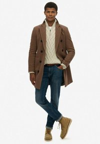 Abrigo de espiga marrón con botones dobles, suéter de punto trenzado crema, jeans azules y botas de cordones color beige. Estilo simple y clásico.