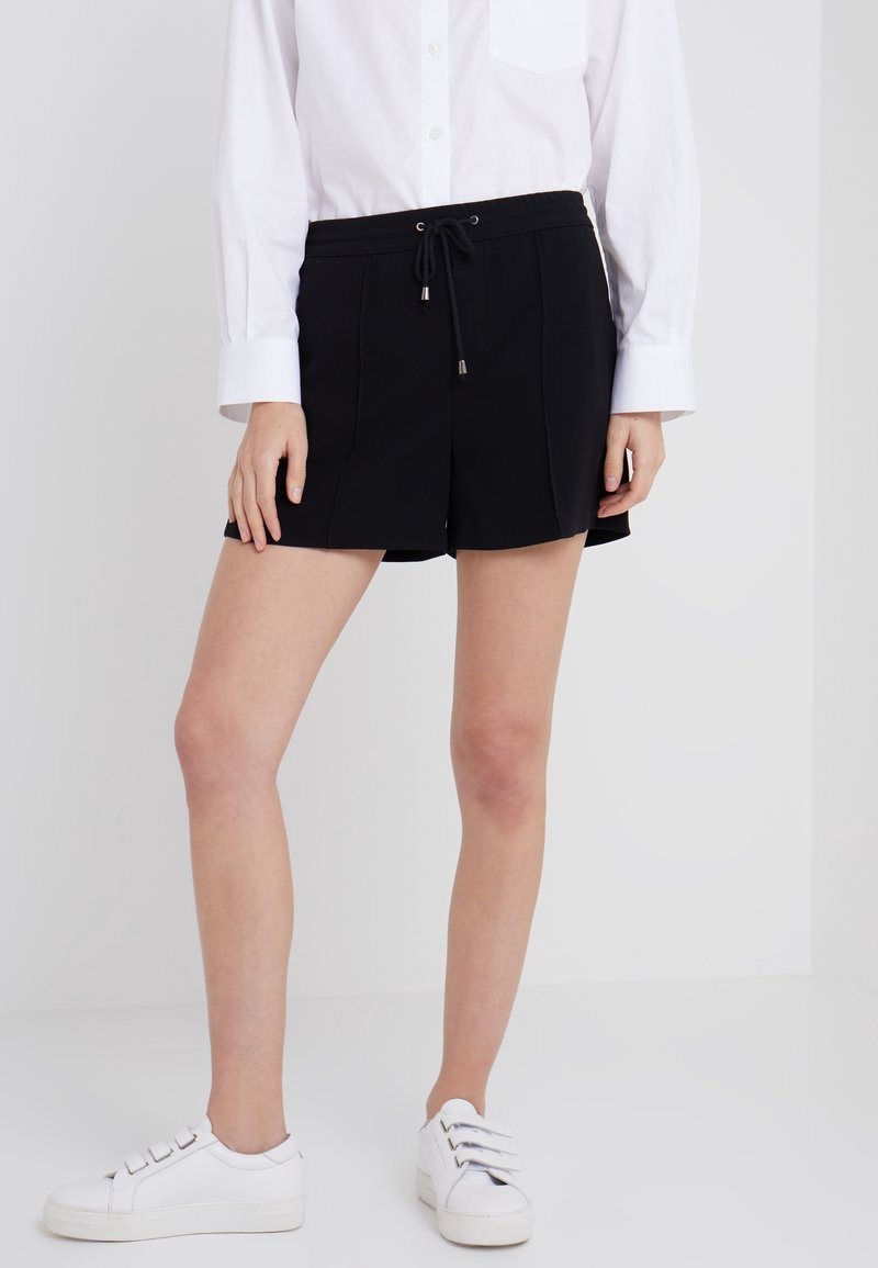 Personne portant un short noir à cordon, une chemise blanche à manches longues et des baskets blanches, debout devant un fond blanc.