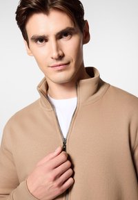 Beżowa bluza rozpinana z wysokim kołnierzem, o gładkiej fakturze i srebrnym zamkiem, noszona na białej koszulce, z ręką trzymającą zamek.