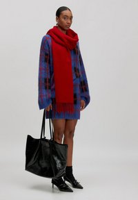 Overmaatse blauwe geruite sweater met rode accenten, gecombineerd met een rode sjaal en een zwarte glanzende handtas. Zwarte puntige hakken maken de look compleet.