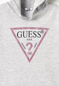 Sudadera gris con capucha y un logo triangular de purpurina rosa que incluye el texto "GUESS" y "U.S.A." junto con detalles de "WASHED" y "JEANS".