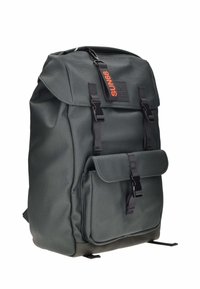 Mochila de tela verde oscuro con la parte superior plegable, que cuenta con correas negras, un bolsillo en la solapa frontal y una etiqueta de logo naranja. Textura suave.