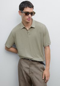 Polo a maniche corte di colore verde oliva con colletto, abbinato a pantaloni marroni dalle texture e occhiali da sole rettangolari scuri. Design semplice e moderno.
