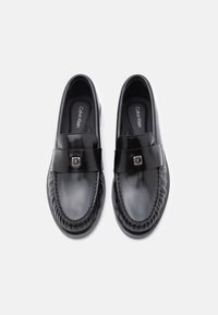 Μαύρα δερμάτινα μοκασίνια με ομαλή υφή, μία λεπτομέρεια με λουράκι στην κορυφή και ασημένιο λογότυπο. Κλασικός σχεδιασμός με ραφές ως διακοσμητικά στοιχεία.