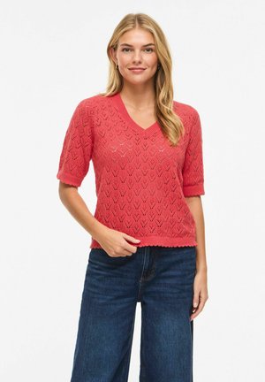 Femme aux longs cheveux blonds portant un pull en maille corail à manches courtes et un jean bleu foncé, debout avec une main touchant l'ourlet du pull.