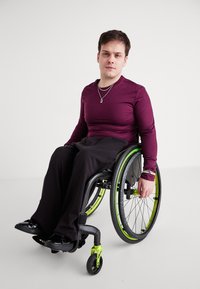 Ένα άτομο σε ένα σκοτεινό αναπηρικό καροτσάκι με πράσινες λεπτομέρειες φοράει ένα μπορντώ μπλ�ουζάκι με μακρυά μανίκια και μαύρο παντελόνι, με σκούρες παπούτσια και κοσμήματα.