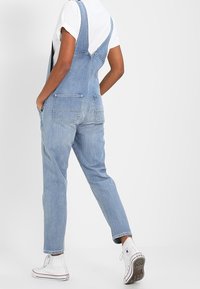 Peto de denim azul con tirantes ajustables, dos bolsillos traseros, corte holgado y dobladillo acampanado, combinado con una camiseta blanca y zapatillas.