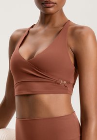 Hnedý športový crop top s výstrihom do V, nastaviteľnými ramienkami, prekrývajúcim dizajnom a zlatými kovovými akcentmi pri bočnom šve. Hladká, pružná tkanina.