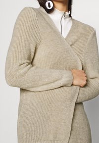 Beige knitted cardigan with a v-neck design, ribbed texture, and long sleeves. Features a relaxed fit and overlapping front panels.