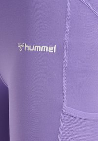 Fioletowe legginsy sportowe marki Hummel, wykonane z gładkiego materiału, z płaskimi szwami i małym białym logo. Zawierają boczną kieszeń dla większej funkcjonalności.