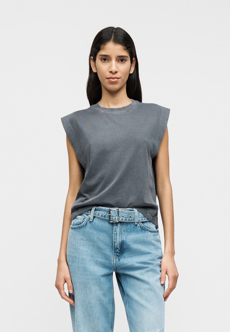 Camiseta sin mangas gris con cuello redondo y corte holgado, combinada con jeans azul claro y un cinturón, mostrada sobre un fondo liso.