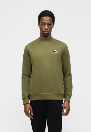 Junger Mann trägt ein olivgrünes Sweatshirt mit Rundhalsausschnitt und kleinem schwarz-weißem Logo auf der linken Brust, kombiniert mit schwarzen Hosen.