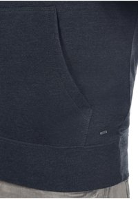 Gros plan sur la poche d'un sweat-shirt gris foncé et le bas du torse d'une personne portant un pantalon clair.