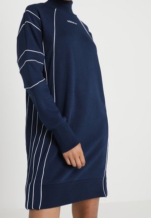 Mørkeblå langærmet kjole med hvide lodrette og skrå striber, høj halsudskæring og lille Adidas-logo på brystet båret af en stående person.
