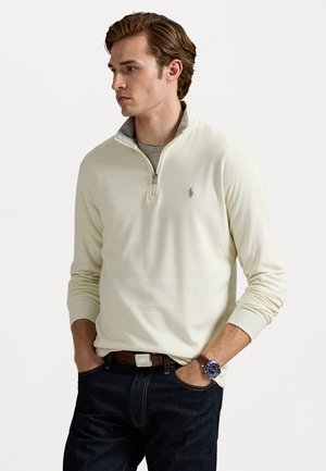 Pulôver de zíper em creme claro com colarinho cinza, apresentando um pequeno logótipo bordado. Usado sobre uma camisa cinza com jeans de denim escuro.