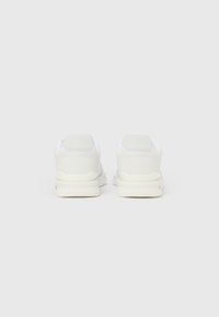 Fehér tornacipő, hálós és szintetikus felsőrésszel, lekerekített sarokkal és texturált díszítésekkel, párnázott talppal és minimalista kialakítással.