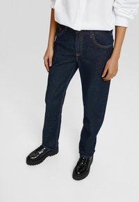 Pantalones de mezclilla azul oscuro con un corte recto, costuras visibles y dos bolsillos frontales, combinados con zapatos negros de cordones.