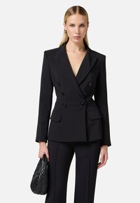 Blazer nero doppiopetto con rever affilati, tasche laterali e bottoni neri, abbinato a pantaloni neri su misura e una borsa in tessuto.