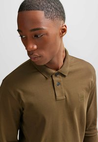 Bruine polo met een kraag, twee blauwe knopen en een klein logopatch. Lange mouwen met een soepele, gestructureerde stof.