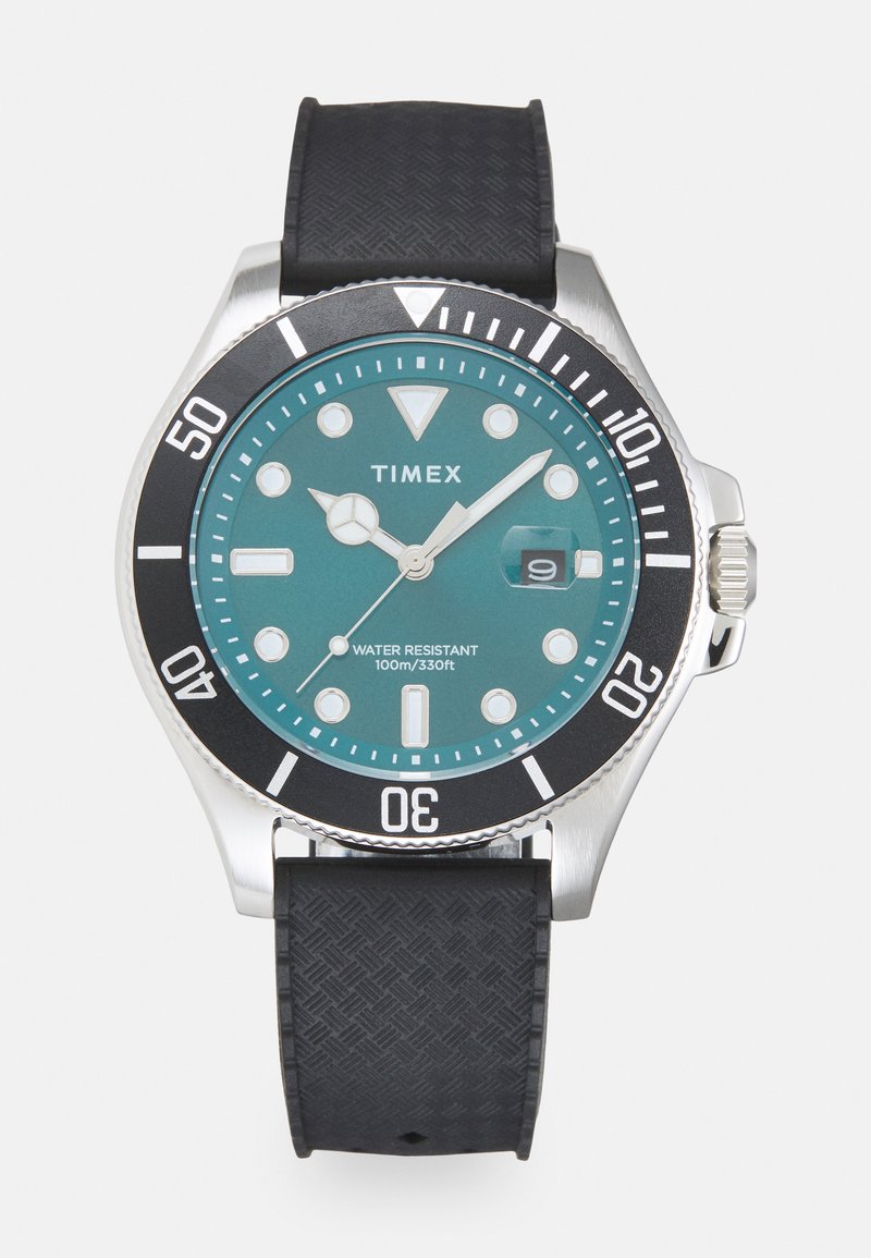 Reloj Timex con una esfera verde azulado, bisel negro y caja plateada. Incluye una correa de goma, marcadores de hora luminosos y una pantalla de fecha a las 3 en punto.