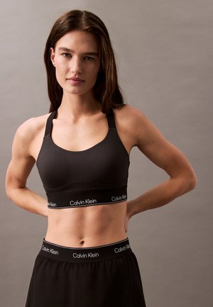 Femme aux longs cheveux bruns portant un soutien-gorge de sport noir Calvin Klein et un short noir Calvin Klein, debout avec les mains derrière le dos.