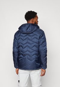 Veste matelassée bleu marine avec capuche, présentant un design en chevrons matelassés et une texture lisse. Manchons et ourlet ajustables pour un meilleur ajustement.