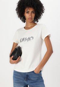 Weißes Baumwoll-T-Shirt mit kurzen Ärmeln, versehen mit silberfarbenem, tonalem Text "LIU JO MILANO" in einer eleganten Schriftart. Kombiniert mit einer kleinen schwarzen Handtasche.
