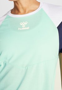 Světle mátová sportovní košile s texturovaným povrchem, bílé akcenty na ramenech a námořnické postranní panely. Obsahuje logo Hummel na hrudi.