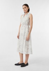 Weißes ärmelloses Kleid mit Kragen und Kordelzug in der Taille, mit einem schwarz gepunkteten Muster. Kombiniert mit schwarzen strukturierten Loafers.