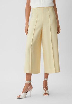 Vrouw draagt beige wijde cropped broek met hoge taille, gecombineerd met witte sandalen met hak en bandjes tegen een effen achtergrond.