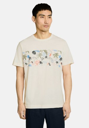 Mężczyzna ubrany w beżowy, krótkim rękawem T-shirt z wielokolorowym tropikalnym wzorem liści i owoców na klatce piersiowej.
