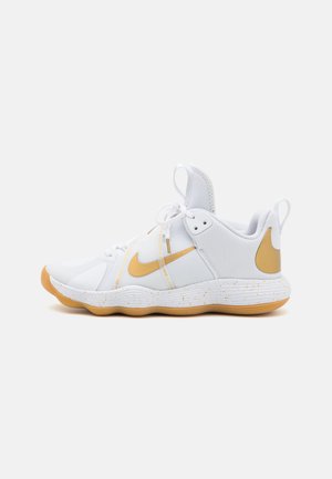 Zapatilla deportiva blanca con parte superior de malla, acentos dorados, entresuela con motas y suela de goma texturizada con punta redondeada.