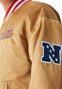 Giacca in satin marrone con colletto a righe rosse e bianche, con la scritta "Raiders" ricamata in rosso e un patch blu con design di una stella bianca.