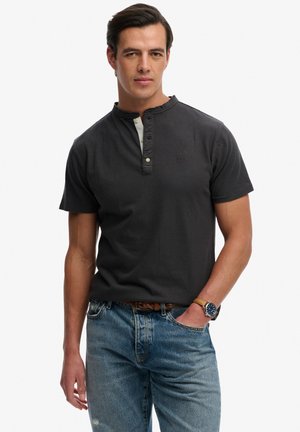 Man draagt een zwart henleyshirt met korte mouwen, blauwe spijkerbroek, bruine riem en een polshorloge, staat met één hand in de zak.