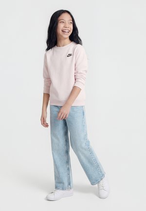 Smilende pige med langt mørkt hår iført en lyserød Nike-sweatshirt, lyseblå jeans og hvide sneakers, stående foran en ensfarvet baggrund.