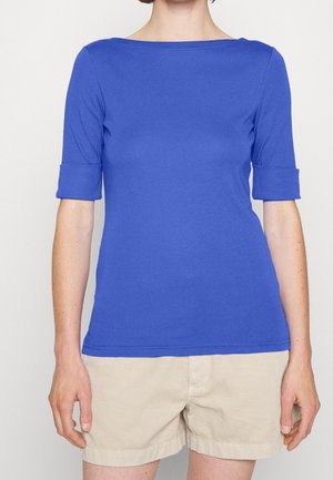 Osoba ubrana w niebieską bluzkę z rękawem do łokcia z podwiniętymi mankietami i beżowe szorty, stojąca na tle jednolitego jasnego tła.