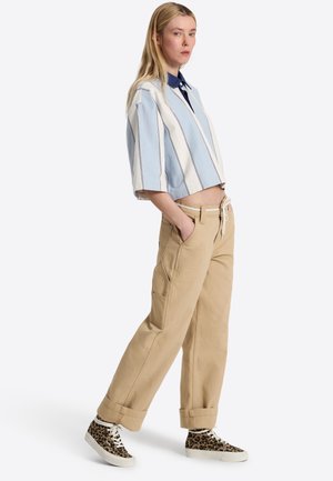 Femme blonde portant une chemise courte rayée bleue et blanche, un pantalon beige à jambes larges, et des baskets montantes à imprimé léopard, debout les mains dans les poches.