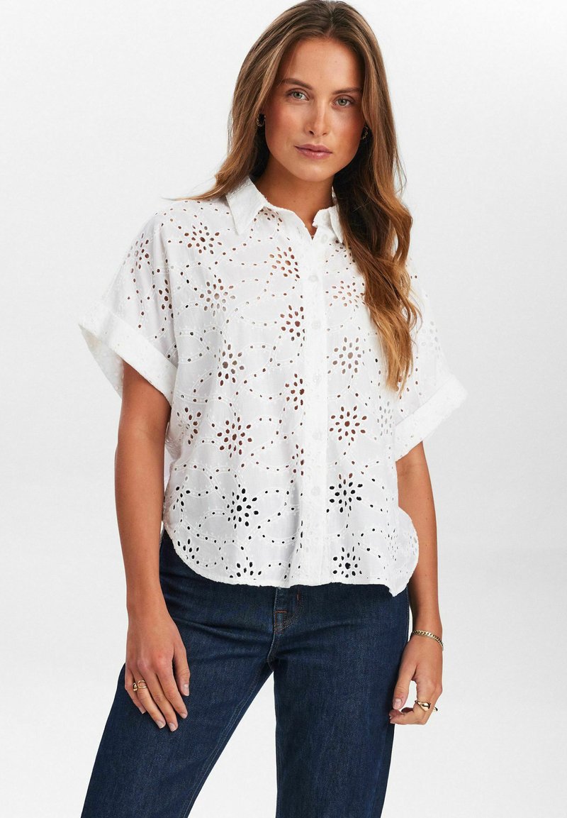 Blusa blanca de manga corta con patrones de bordado de ojal, cierre frontal con botones y un corte relajado, combinada con jeans azul oscuro.