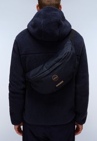 Námořnická taška přes rameno vyrobená z odolné tkaniny, s zakřiveným tvarem, zipovým uzávěrem a výraznou nášivkou loga na přední straně, nošena přes fleecovou bundu.