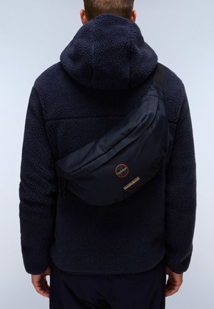 Marineblaues Umhängetasche aus strapazierfähigem Stoff, mit geschwungener Form, Reißverschluss und auffälligem Logo-Patch auf der Vorderseite, getragen über einer Fleecejacke.