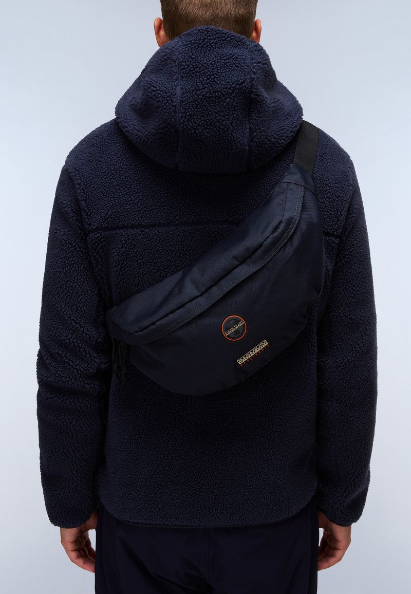 Námořnická taška přes rameno vyrobená z odolné tkaniny, s zakřiveným tvarem, zipovým uzávěrem a výraznou nášivkou loga na přední straně, nošena přes fleecovou bundu.