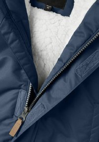 Námořnická modrá bunda s měkkou bílou fleecovou podšívkou, s kovovým zipem, textilními nášivkami a koženým táhlem na zip. Detailní pohled na oblast límce.