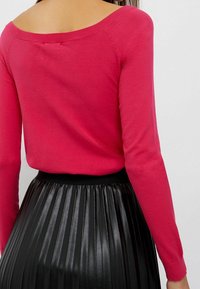 Maglione rosa fitted, con spalle scoperte, realizzato in morbido materiale lavorato a maglia, abbinato a una gonna nera plissettata in similpelle con una texture liscia.