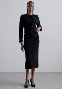 Costum din tricot negru în două piese, format dintr-o jachetă scurtă și o fustă lungă. Prezintă un material texturat și accente cu nasturi pe ambele piese.