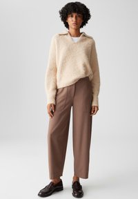 Maglione beige fluffy con colletto, abbinato a pantaloni marroni a vita alta con tasche anteriori e gamba dritta. Scarpe scure completano il look.
