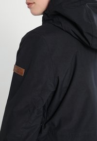 Zwarte jas met capuchon, met een gestructureerd bruin leren patch op de schouder en een gladde stofafwerking. Inclusief ritszakdetails.