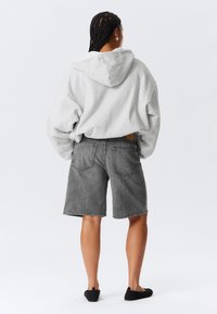 Siva oversize majica s kapuljačom s izvlačnim vrpcom, uparena s sivim traper šortsevima. Šortsevi imaju dva stražnja džepa i detalj istrošenog ruba.