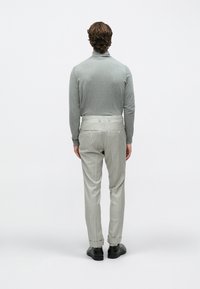 Mand stående med ryggen til iført en grå rullekrave, lysegrå bukser med nålestriber og sorte pæne sko mod en hvid baggrund.