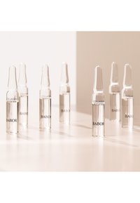 Ampollas de vidrio transparente etiquetadas "AUMENTADOR DE COLÁGENO" y "BABOR", dispuestas sobre una superficie clara, mostrando un diseño minimalista y elegante.